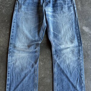 May include: Faded blue denim jeans with a classic five-pocket design. The jeans have a straight leg cut and feature brown leather accents on the front pockets. The denim has a worn, distressed look.