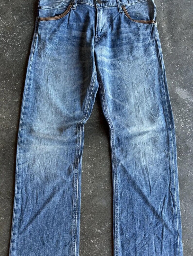 May include: Faded blue denim jeans with a classic five-pocket design. The jeans have a straight leg cut and feature brown leather accents on the front pockets. The denim has a worn, distressed look.