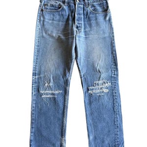 May include: Light blue denim jeans with distressed details, including rips at the knees. The jeans have a button fly, classic five-pocket design, and a faded wash. The straight-leg style is visible.