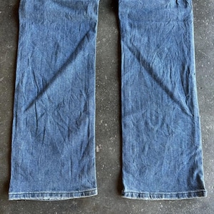 May include: A pair of blue denim jeans. The jeans have a classic five-pocket design, a straight leg, and a medium wash. The fabric shows subtle fading and creasing, suggesting wear. The jeans are laid flat on a textured surface.
