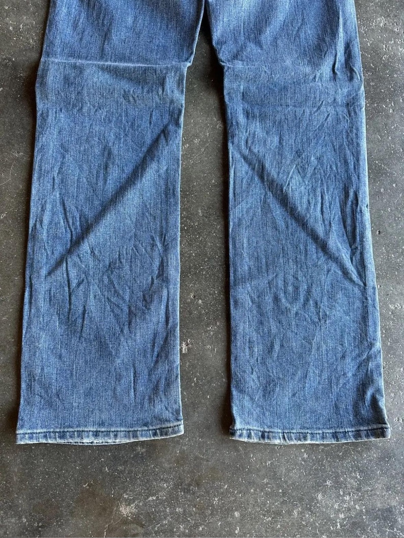 May include: A pair of blue denim jeans. The jeans have a classic five-pocket design, a straight leg, and a medium wash. The fabric shows subtle fading and creasing, suggesting wear. The jeans are laid flat on a textured surface.