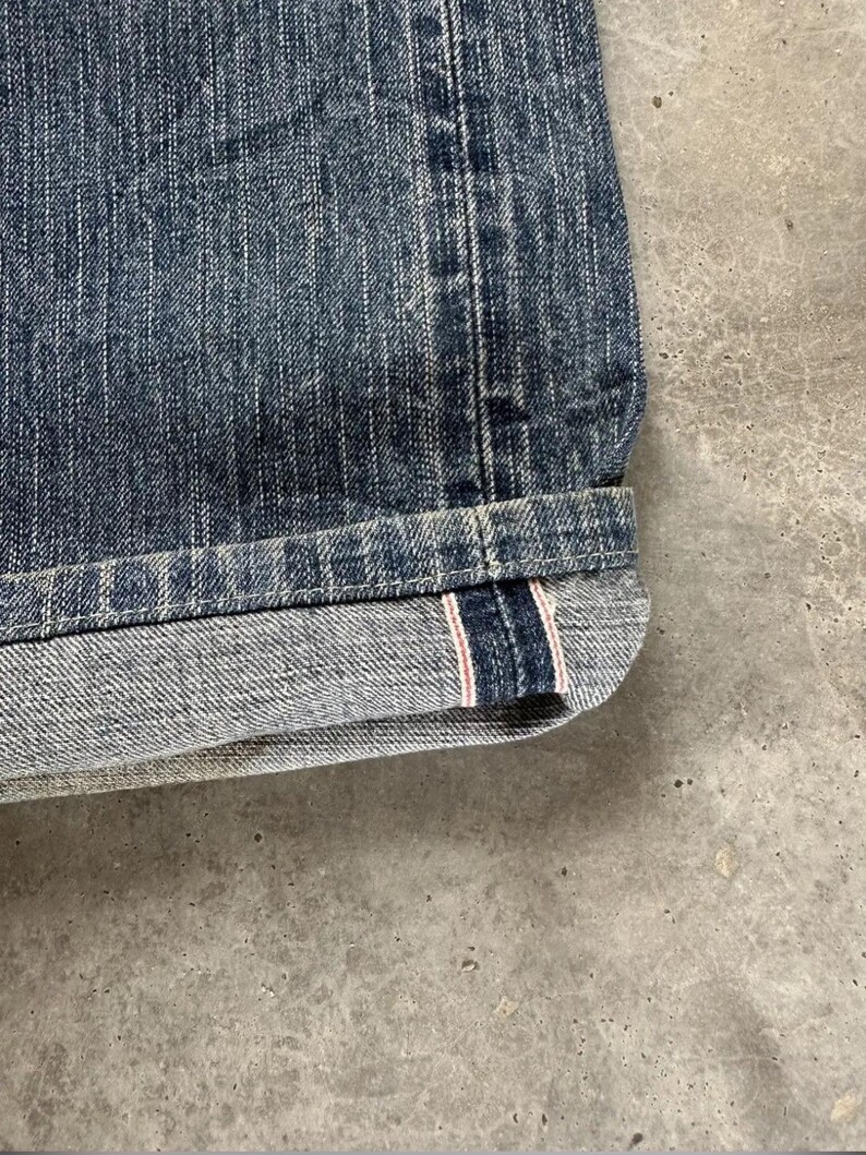 May include: Close-up of the hem of blue denim shorts. The denim has a textured appearance with vertical lines. The hem is cuffed, revealing a red and white stripe detail on the inside.