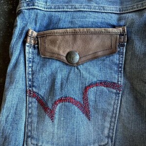 May include: Close-up of light blue denim jeans with a brown leather pocket and a red embroidered design on the back pocket. The pocket has a silver button with the text "BLUE TRIP".