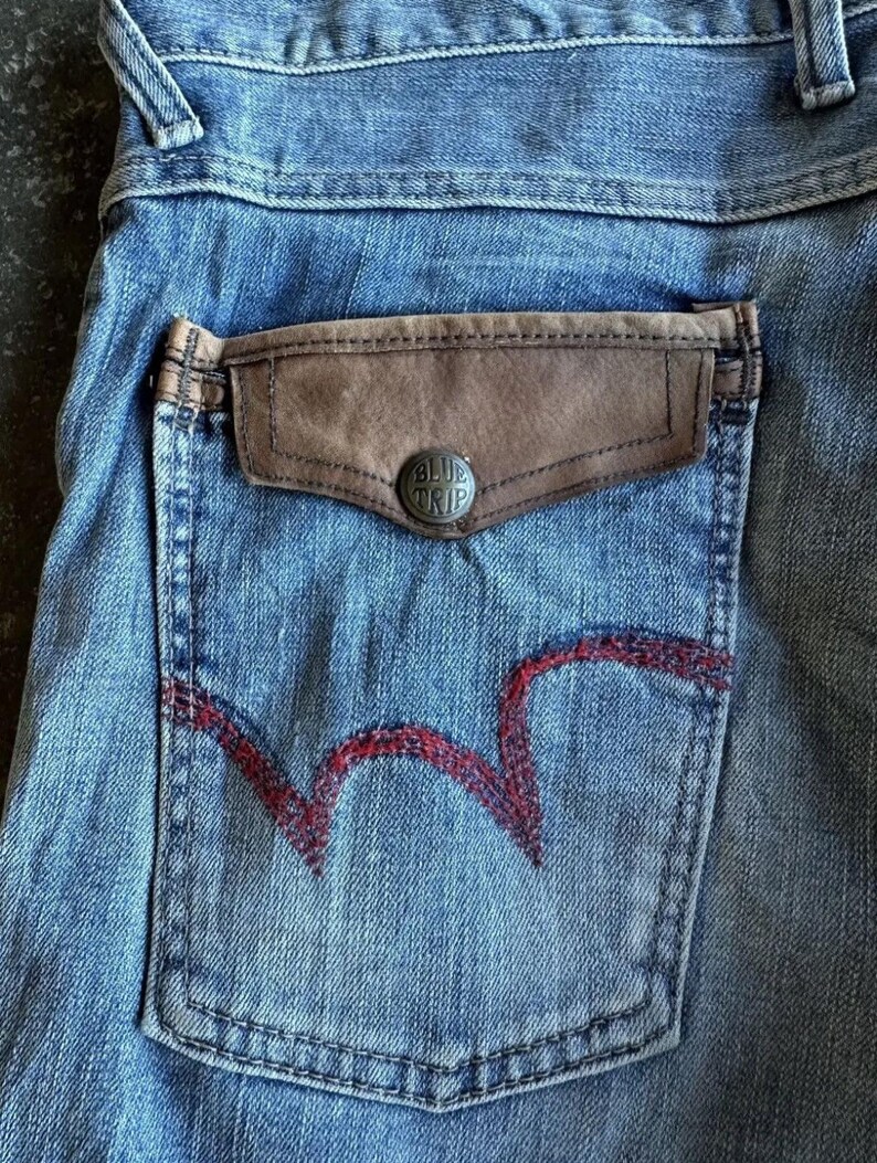 May include: Close-up of light blue denim jeans with a brown leather pocket and a red embroidered design on the back pocket. The pocket has a silver button with the text "BLUE TRIP".