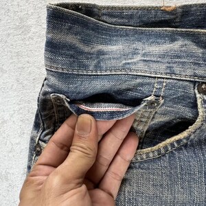 May include: Close-up of blue denim jeans with a distressed wash. The jeans feature a classic five-pocket design, with visible stitching and rivets. The pocket lining has a pink accent. The denim has a worn, vintage look.