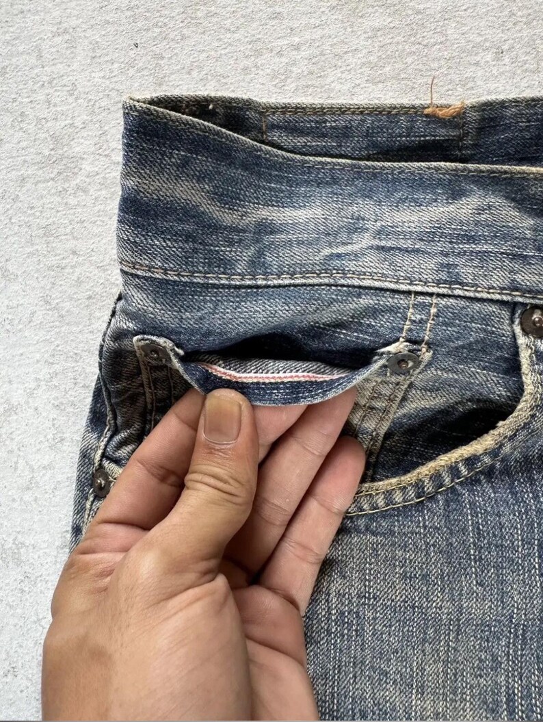 May include: Close-up of blue denim jeans with a distressed wash. The jeans feature a classic five-pocket design, with visible stitching and rivets. The pocket lining has a pink accent. The denim has a worn, vintage look.