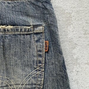 May include: Close-up of blue denim jeans with a distressed appearance. The jeans have a back pocket with a brown leather tag that reads "EDWIN". White stitching details the pocket and the seams of the jeans.