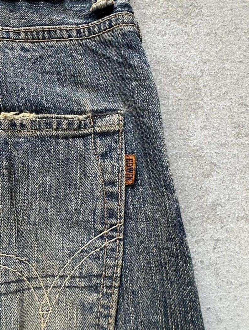 May include: Close-up of blue denim jeans with a distressed appearance. The jeans have a back pocket with a brown leather tag that reads "EDWIN". White stitching details the pocket and the seams of the jeans.
