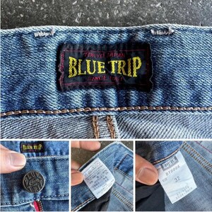 May include: Close-up of blue denim jeans with a "BLUE TRIP" label. The label is black with yellow text. Additional details include a button with "BLUE TRIP" and a size 32 tag.
