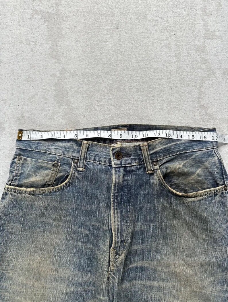 May include: Close-up of blue denim jeans with a tape measure across the waistband. The jeans show signs of wear and fading. The tape measure indicates a waist measurement of approximately 45.7 cm.