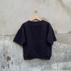 May include: A black short-sleeved sweatshirt hanging on a wooden hanger against a gray concrete wall. The sweatshirt has a crew neck and a relaxed fit. The sleeves are short and wide. The garment appears to be made of a soft, thick material.