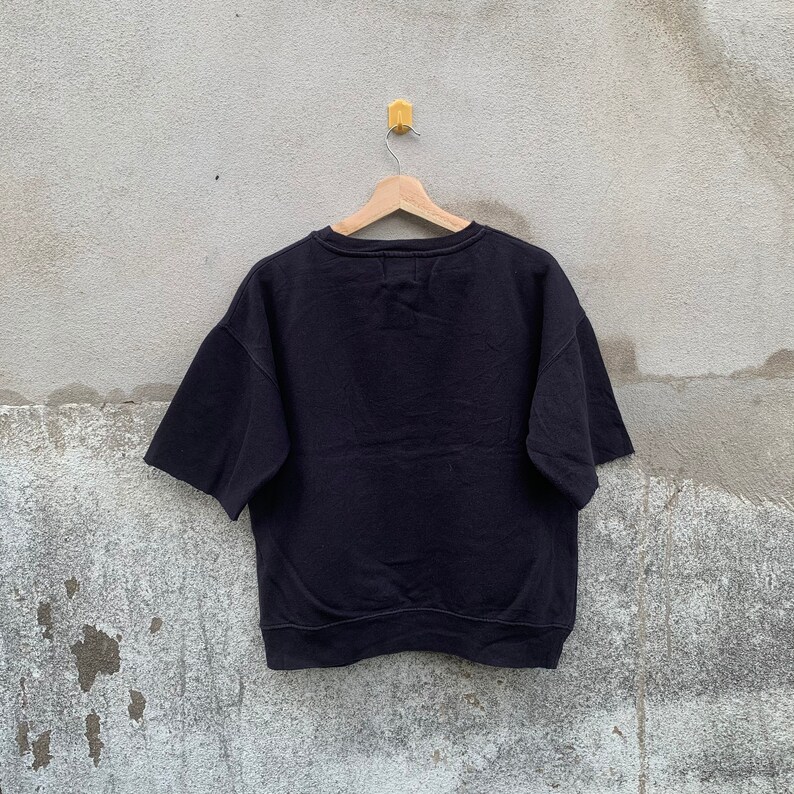 May include: A black short-sleeved sweatshirt hanging on a wooden hanger against a gray concrete wall. The sweatshirt has a crew neck and a relaxed fit. The sleeves are short and wide. The garment appears to be made of a soft, thick material.