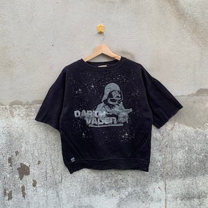 May include: Black short-sleeved sweatshirt featuring a gray graphic of Darth Vader from Star Wars. The graphic includes the text "Darth Vader" and a starry background. The sweatshirt is hanging on a wooden hanger.