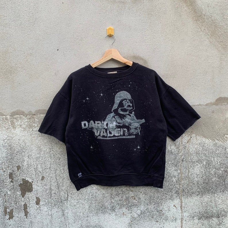 May include: Black short-sleeved sweatshirt featuring a gray graphic of Darth Vader from Star Wars. The graphic includes the text "Darth Vader" and a starry background. The sweatshirt is hanging on a wooden hanger.