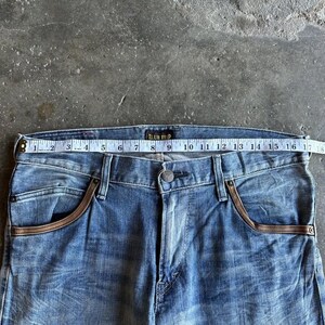 May include: Light blue denim jeans with brown pocket trim. The jeans are measured with a tape measure, showing a waist size of approximately 17 inches. The brand name "BLUE TRIP" is visible on the waistband.