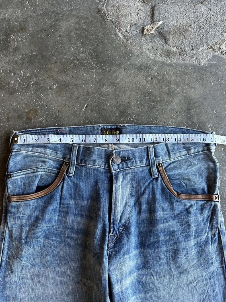 May include: Light blue denim jeans with brown pocket trim. The jeans are measured with a tape measure, showing a waist size of approximately 17 inches. The brand name "BLUE TRIP" is visible on the waistband.