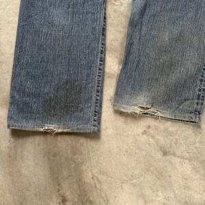 May include: Close-up of the lower section of a pair of worn blue denim jeans. The jeans show signs of wear and tear, with frayed edges at the bottom hems. The denim has faded patches and darker areas, indicating age and use. The jeans are laid flat on a concrete surface.