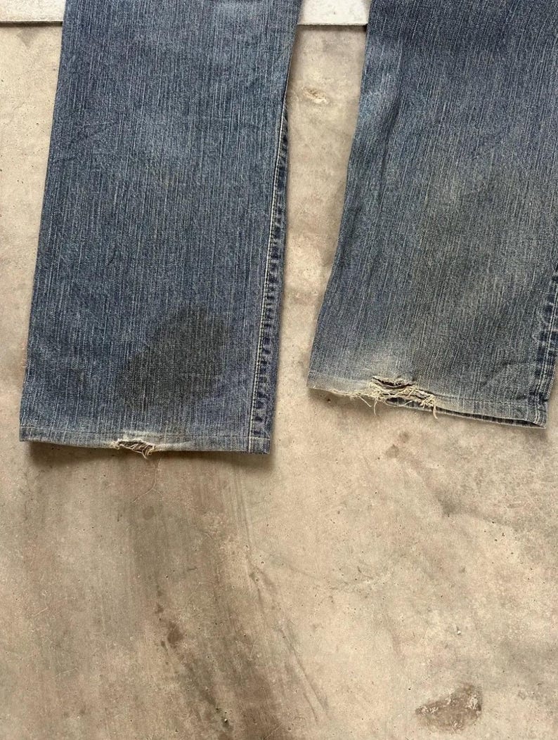 May include: Close-up of the lower section of a pair of worn blue denim jeans. The jeans show signs of wear and tear, with frayed edges at the bottom hems. The denim has faded patches and darker areas, indicating age and use. The jeans are laid flat on a concrete surface.