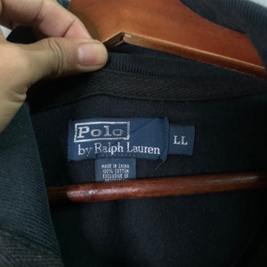 May include: Close-up of a black Polo by Ralph Lauren label. The label is navy blue with white text and the brand logo. The size is LL. The tag reads "Made in China 100% Cotton Exclusive of Decoration."