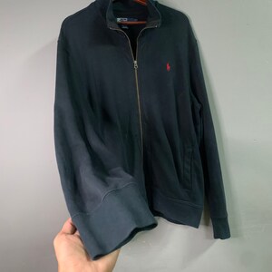 May include: A navy blue zip-up sweatshirt with a small red logo on the chest. The sweatshirt has a full zipper, a high collar, and ribbed cuffs and hem. The brand name is visible on the collar.