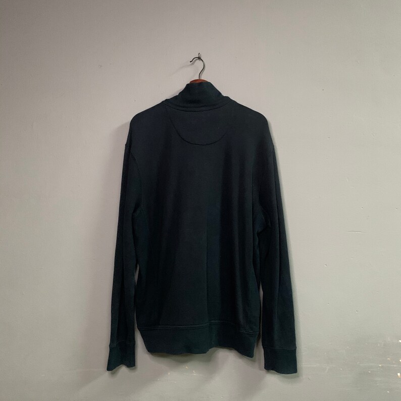 May include: A dark teal sweatshirt with a mock neck collar and long sleeves. The back of the sweatshirt is visible, hanging from a metal hook. The garment has a simple, classic design, suitable for casual wear.