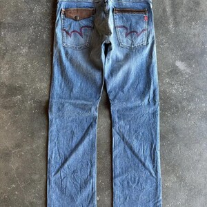 May include: Light blue denim jeans with brown leather pocket accents and red stitching on the back pockets. The jeans have a straight leg cut and a leather patch on the waistband. The jeans are laid flat on a concrete surface.