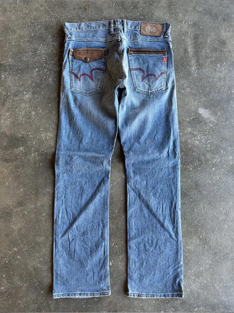 May include: Light blue denim jeans with brown leather pocket accents and red stitching on the back pockets. The jeans have a straight leg cut and a leather patch on the waistband. The jeans are laid flat on a concrete surface.