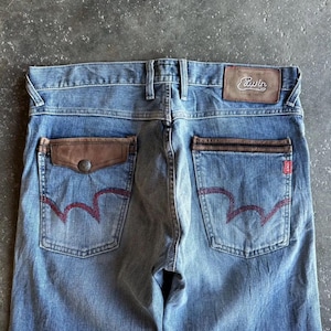 May include: Faded blue denim jeans with brown leather pocket accents and a leather patch with the brand name "Edwin". The back pockets feature a distinctive red stitching design. The jeans have a classic five-pocket design.