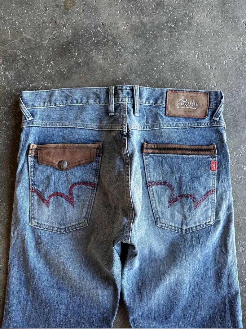 May include: Faded blue denim jeans with brown leather pocket accents and a leather patch with the brand name "Edwin". The back pockets feature a distinctive red stitching design. The jeans have a classic five-pocket design.