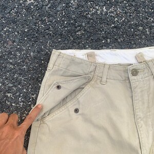 May include: Close-up of khaki-colored pants with a unique pocket design. The pants feature a button closure and a distinctive pocket flap secured by metal snaps. The fabric appears to be a durable cotton blend, suitable for casual wear.