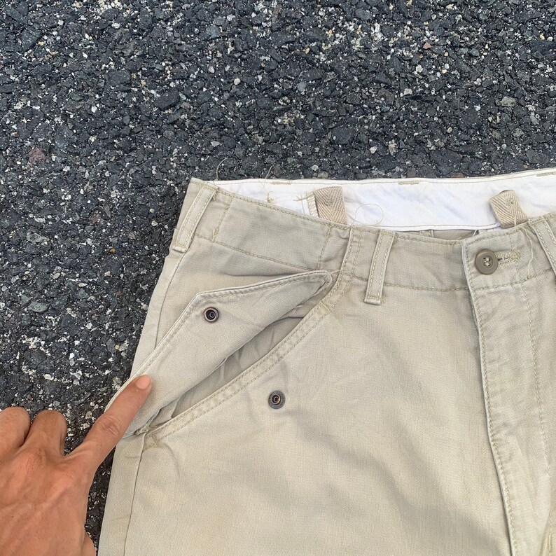 May include: Close-up of khaki-colored pants with a unique pocket design. The pants feature a button closure and a distinctive pocket flap secured by metal snaps. The fabric appears to be a durable cotton blend, suitable for casual wear.