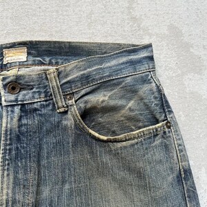 May include: Close-up of faded blue denim jeans, showing wear and tear with visible fading and distressing. The pocket and button details are prominent, showcasing the texture and colour variations of the denim fabric. The tag reads "Made in Japan".