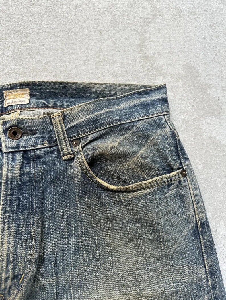 May include: Close-up of faded blue denim jeans, showing wear and tear with visible fading and distressing. The pocket and button details are prominent, showcasing the texture and colour variations of the denim fabric. The tag reads "Made in Japan".