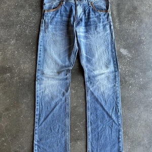 May include: Faded blue denim jeans with a classic five-pocket design. The jeans have a straight leg cut and show signs of wear, with lighter areas on the thighs and knees. The pockets have a brown trim. The jeans are laid flat on a gray surface.