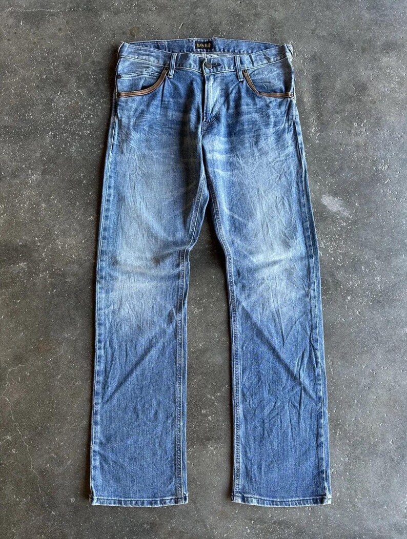 May include: Faded blue denim jeans with a classic five-pocket design. The jeans have a straight leg cut and show signs of wear, with lighter areas on the thighs and knees. The pockets have a brown trim. The jeans are laid flat on a gray surface.