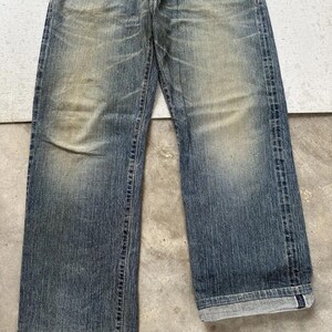 May include: Faded blue denim jeans with a straight leg cut. The jeans show signs of wear with lighter patches and subtle distressing. The cuffs are rolled up, revealing the interior fabric. The jeans are a classic style.