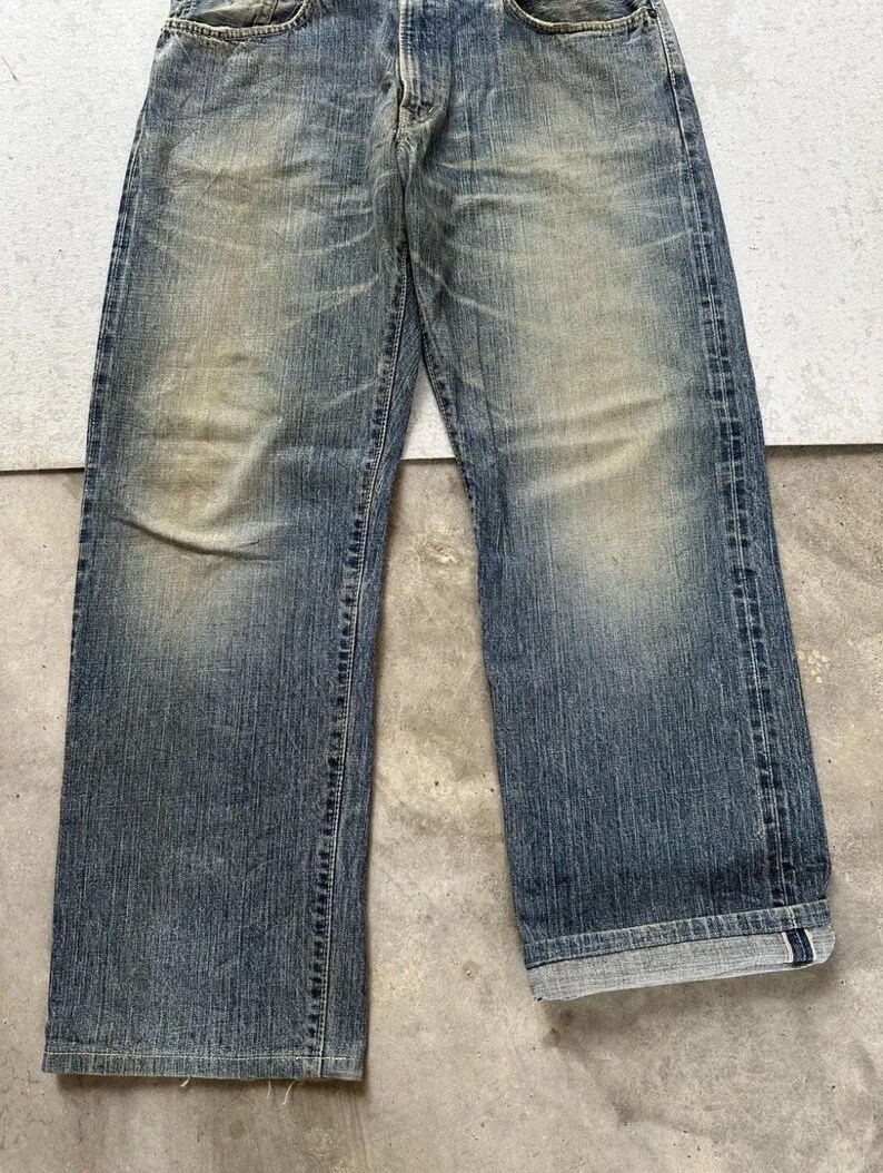 May include: Faded blue denim jeans with a straight leg cut. The jeans show signs of wear with lighter patches and subtle distressing. The cuffs are rolled up, revealing the interior fabric. The jeans are a classic style.