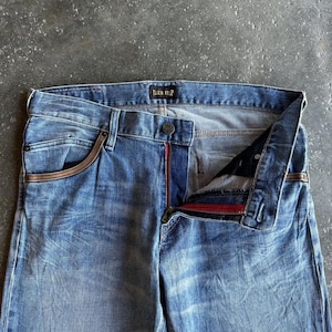 May include: Light blue denim jeans with a faded wash. The jeans have a button closure, a red zipper, and brown accents on the pockets. The brand name "BLUE TRIP" is visible on the waistband.