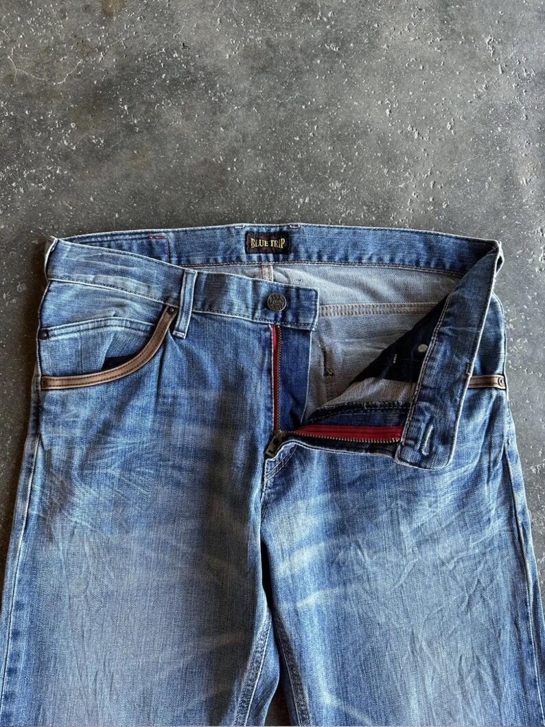 May include: Light blue denim jeans with a faded wash. The jeans have a button closure, a red zipper, and brown accents on the pockets. The brand name "BLUE TRIP" is visible on the waistband.