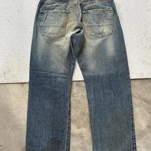 May include: Faded blue denim jeans with a distressed appearance. The jeans feature a classic five-pocket design, a leather patch, and a worn look. The jeans are a straight-leg style.