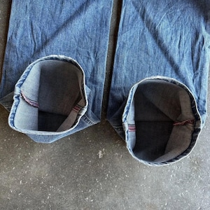 May include: Close-up of blue denim jeans with the cuffs rolled up, revealing the inside lining. The jeans are a medium wash and appear worn. The cuffs show a red seam.