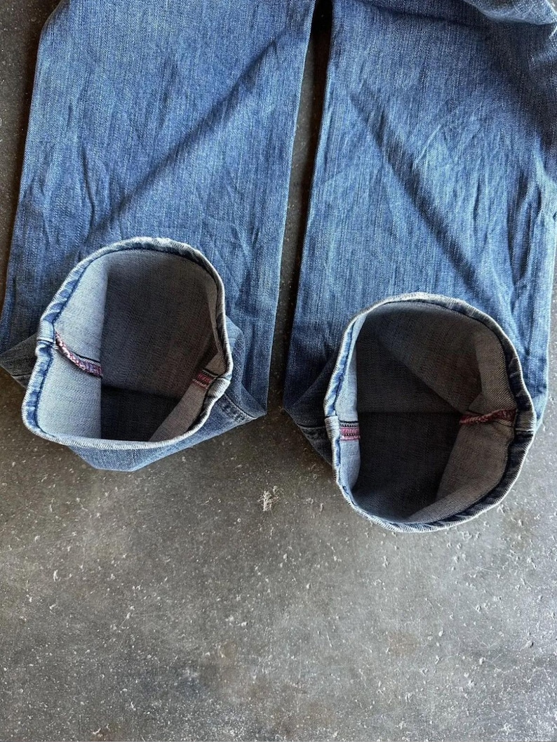 May include: Close-up of blue denim jeans with the cuffs rolled up, revealing the inside lining. The jeans are a medium wash and appear worn. The cuffs show a red seam.