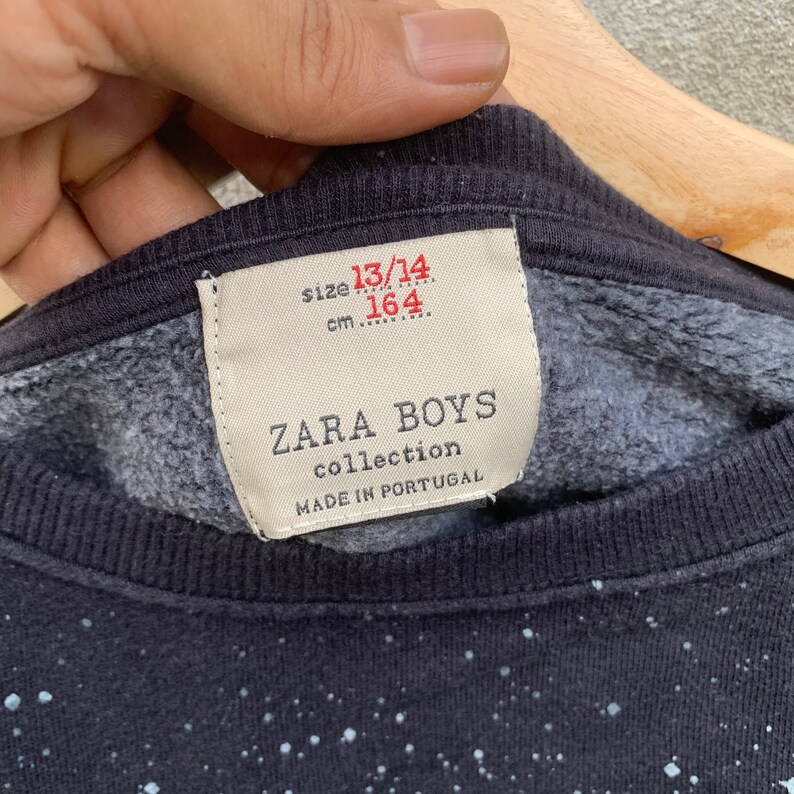 May include: Close-up of a navy blue sweatshirt with a speckled pattern. The label reads "ZARA BOYS collection MADE IN PORTUGAL" with size 13/14 and 164 cm. The sweatshirt is held by a hand.