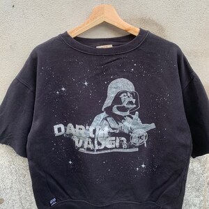May include: Black short-sleeved sweatshirt featuring a graphic of Darth Vader with the text "Darth Vader" in a distressed style. The design includes a starry background. The sweatshirt is hanging on a wooden hanger.
