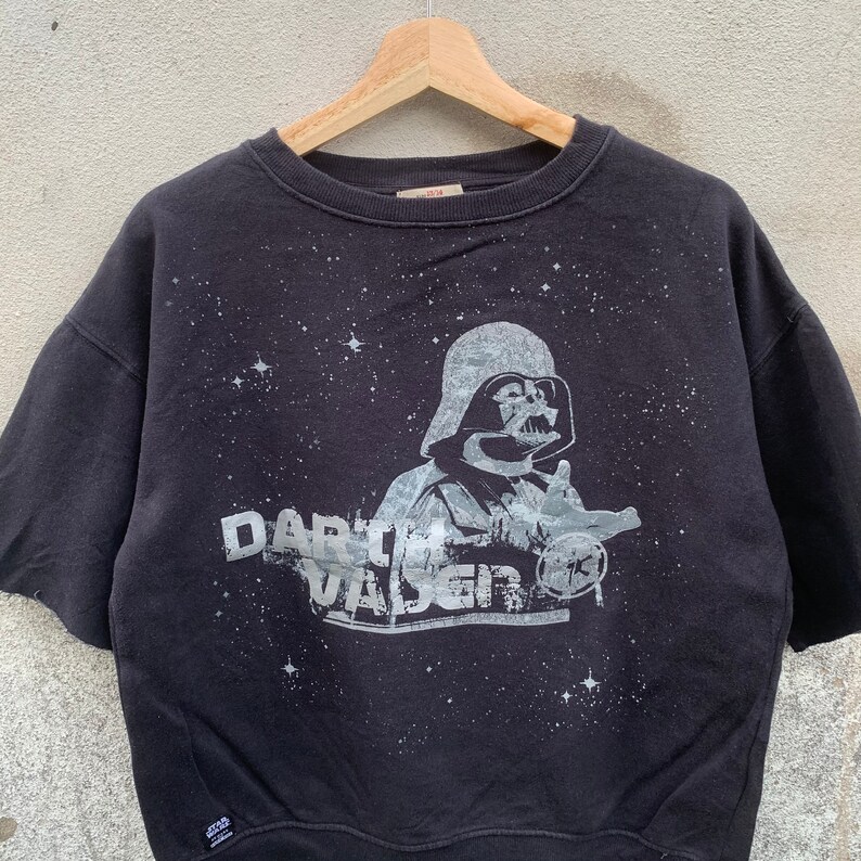 May include: Black short-sleeved sweatshirt featuring a graphic of Darth Vader with the text "Darth Vader" in a distressed style. The design includes a starry background. The sweatshirt is hanging on a wooden hanger.