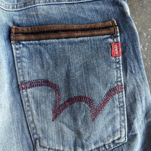 May include: Close-up of light blue denim jeans with a brown leather patch and a red tag. The back pocket features a distinctive red stitched design. The jeans have a classic, worn look.