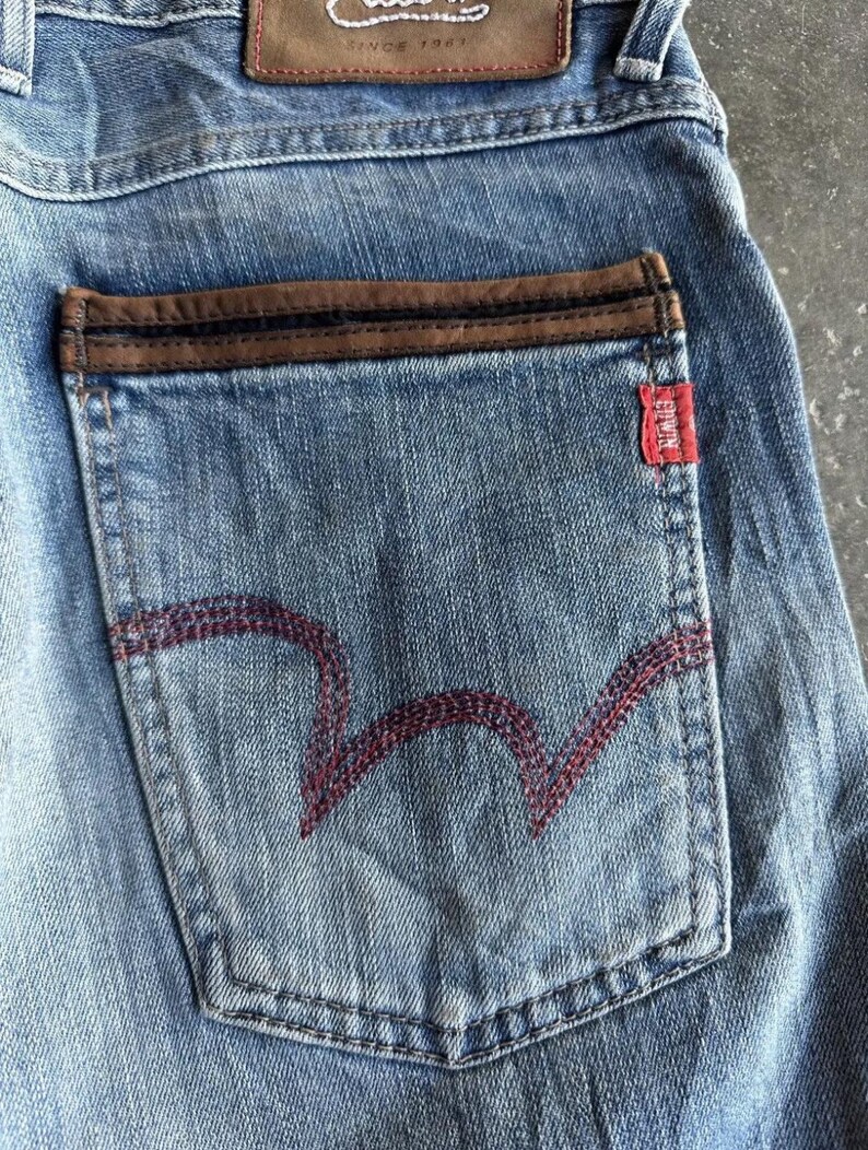 May include: Close-up of light blue denim jeans with a brown leather patch and a red tag. The back pocket features a distinctive red stitched design. The jeans have a classic, worn look.