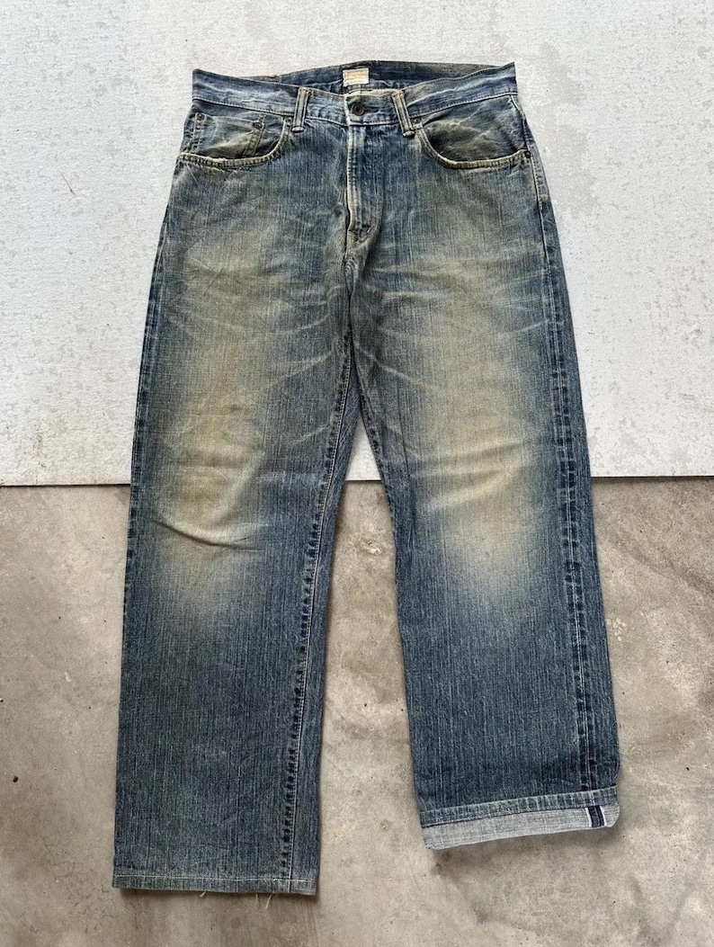 May include: Faded blue denim jeans with a straight leg cut. The jeans show signs of wear and tear, with lighter patches and a worn-in look. Classic five-pocket design with a button and zip closure.