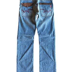 May include: Light blue denim jeans with a faded wash. The back pockets feature a unique red stitching design and brown leather accents. The jeans have a classic five-pocket design and a straight leg fit.