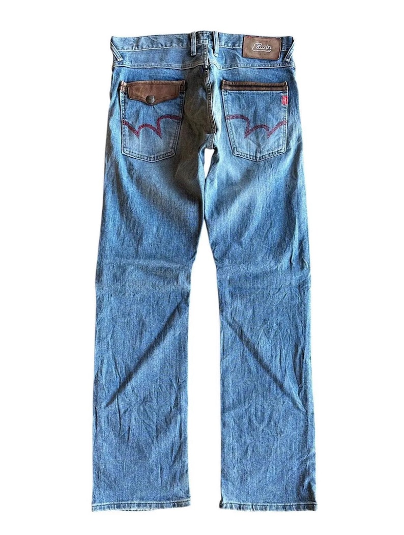 May include: Light blue denim jeans with a faded wash. The back pockets feature a unique red stitching design and brown leather accents. The jeans have a classic five-pocket design and a straight leg fit.